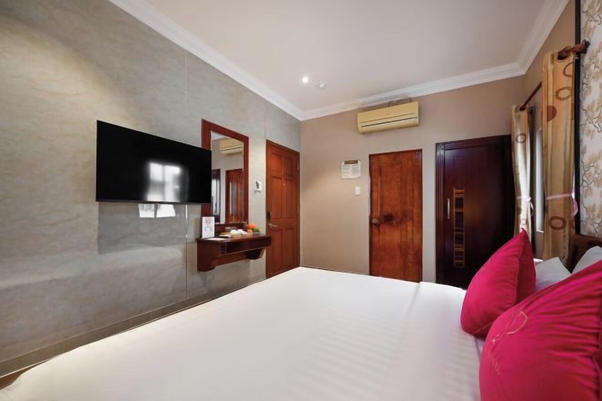 A hotel room with a large bed, a flat-screen TV mounted on the wall, a wooden wardrobe, and an air conditioning unit.