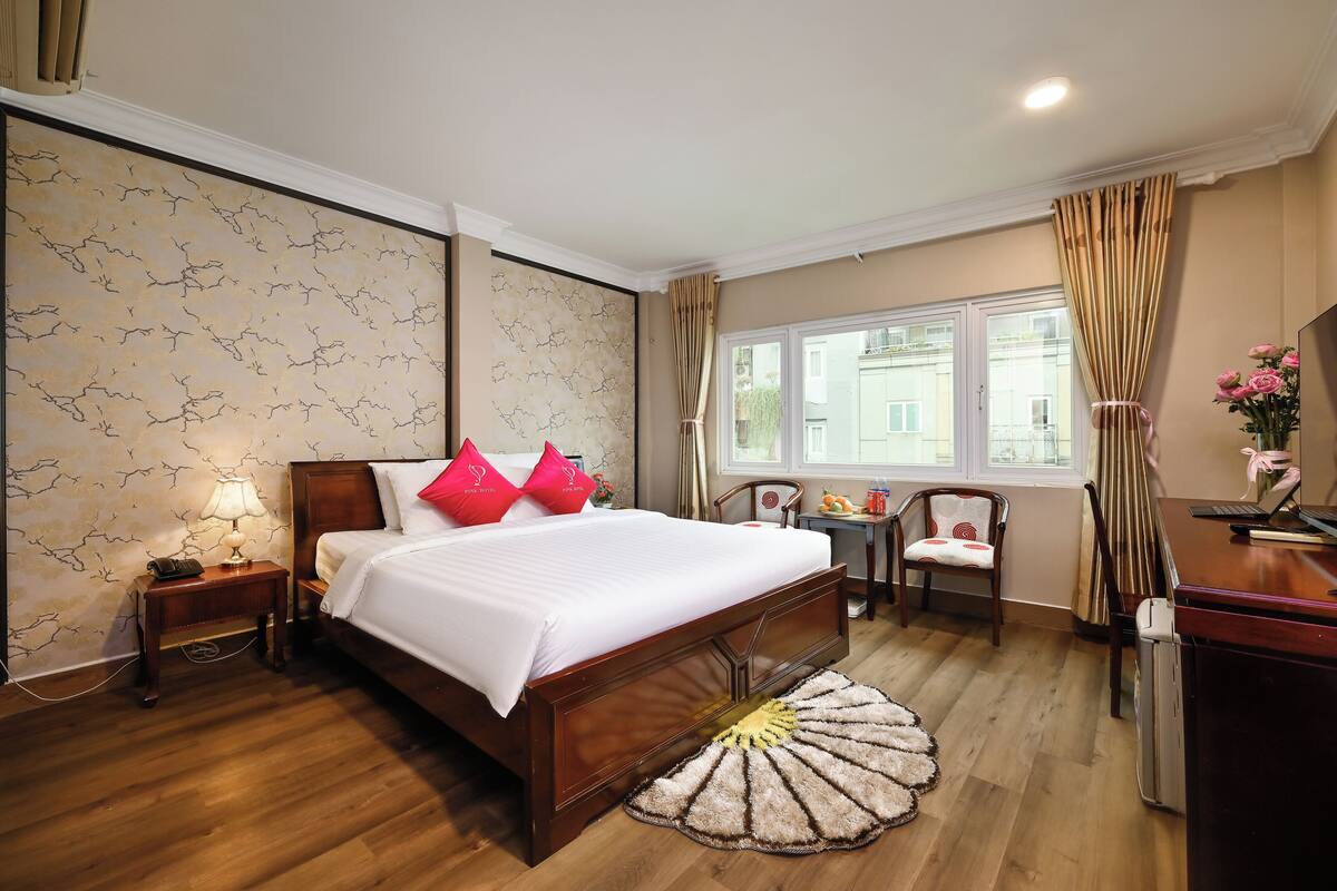 Quarto de hotel com cama de madeira, mesa de cabeceira com luminária, pequena mesa com cadeira e janela com cortinas.