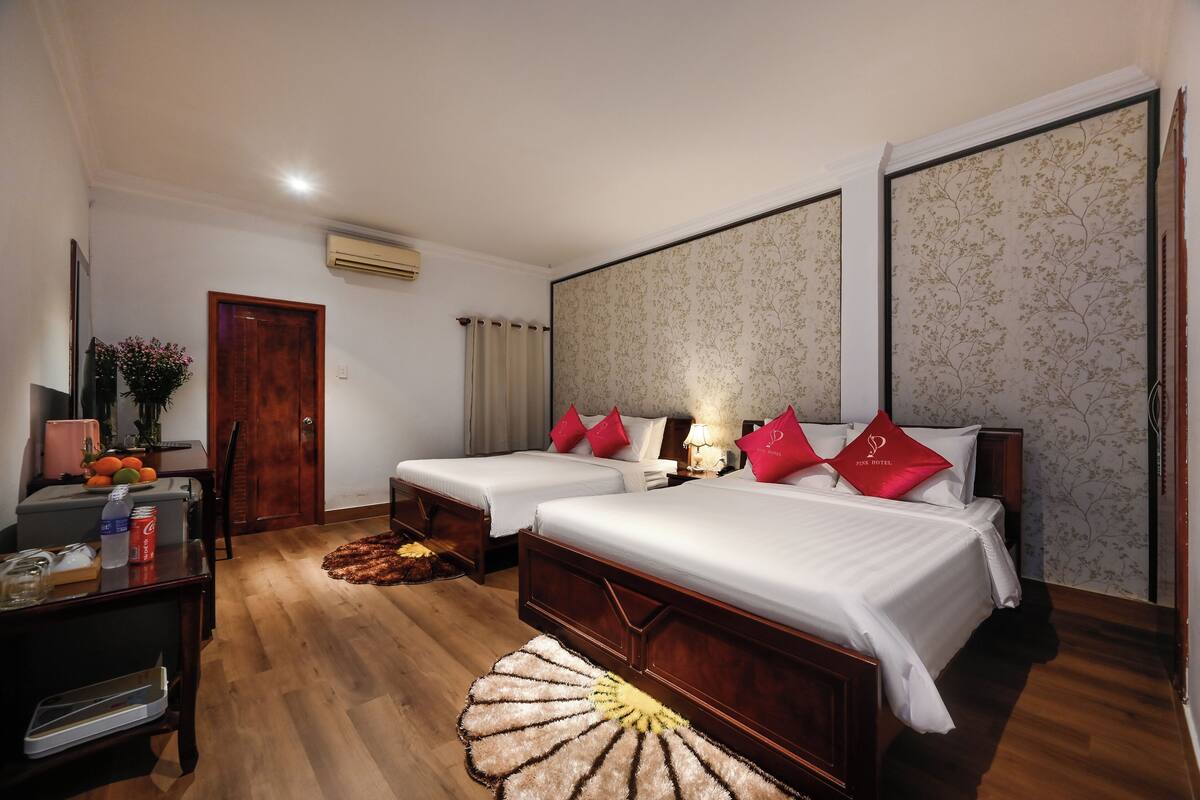 Quarto de hotel com duas camas, uma secretária com um computador, uma cadeira, uma pequena mesa com frutas e um tapete decorativo.