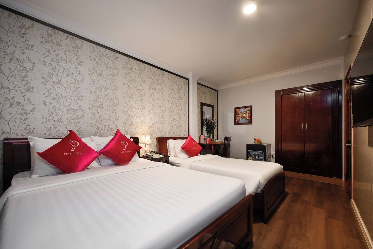 Quarto de hotel com duas camas, um guarda-roupa de madeira, uma escrivaninha com cadeira, um espelho e um quadro na parede.