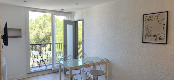 A room with a glass table and chairs, a door leading to a balcony with outdoor furniture, and a framed abstract art piece on the wall.