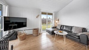 A modern living room with a large flat-screen TV, a leather sofa, a small round table, and a wicker basket. - Charming holiday home close to the North Sea and Søndervig center (Ringkøbing)