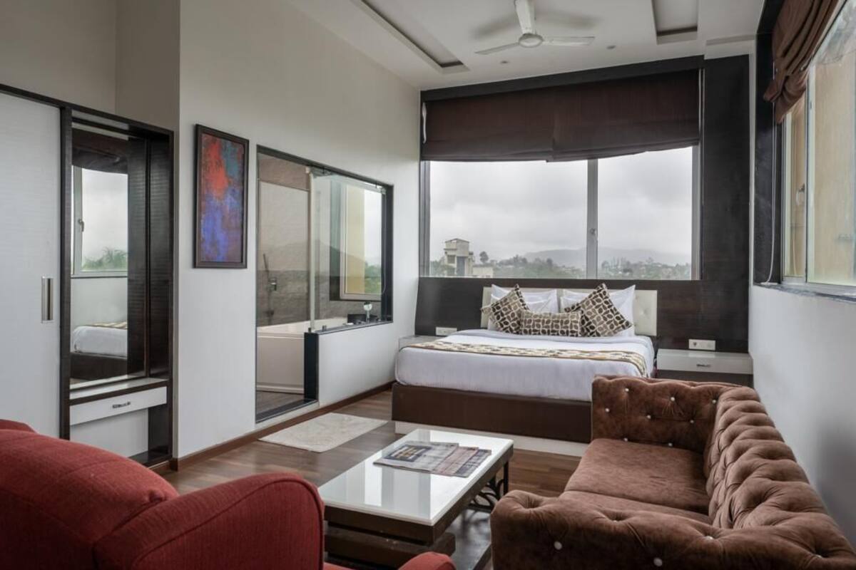 A modern bedroom with a large bed, a sofa, a coffee table, and a view of the city through large windows.