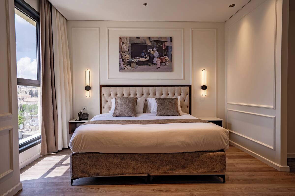 A large bed with a tufted headboard, two bedside tables, a framed picture above the bed, and a view of the city through a window with curtains.
