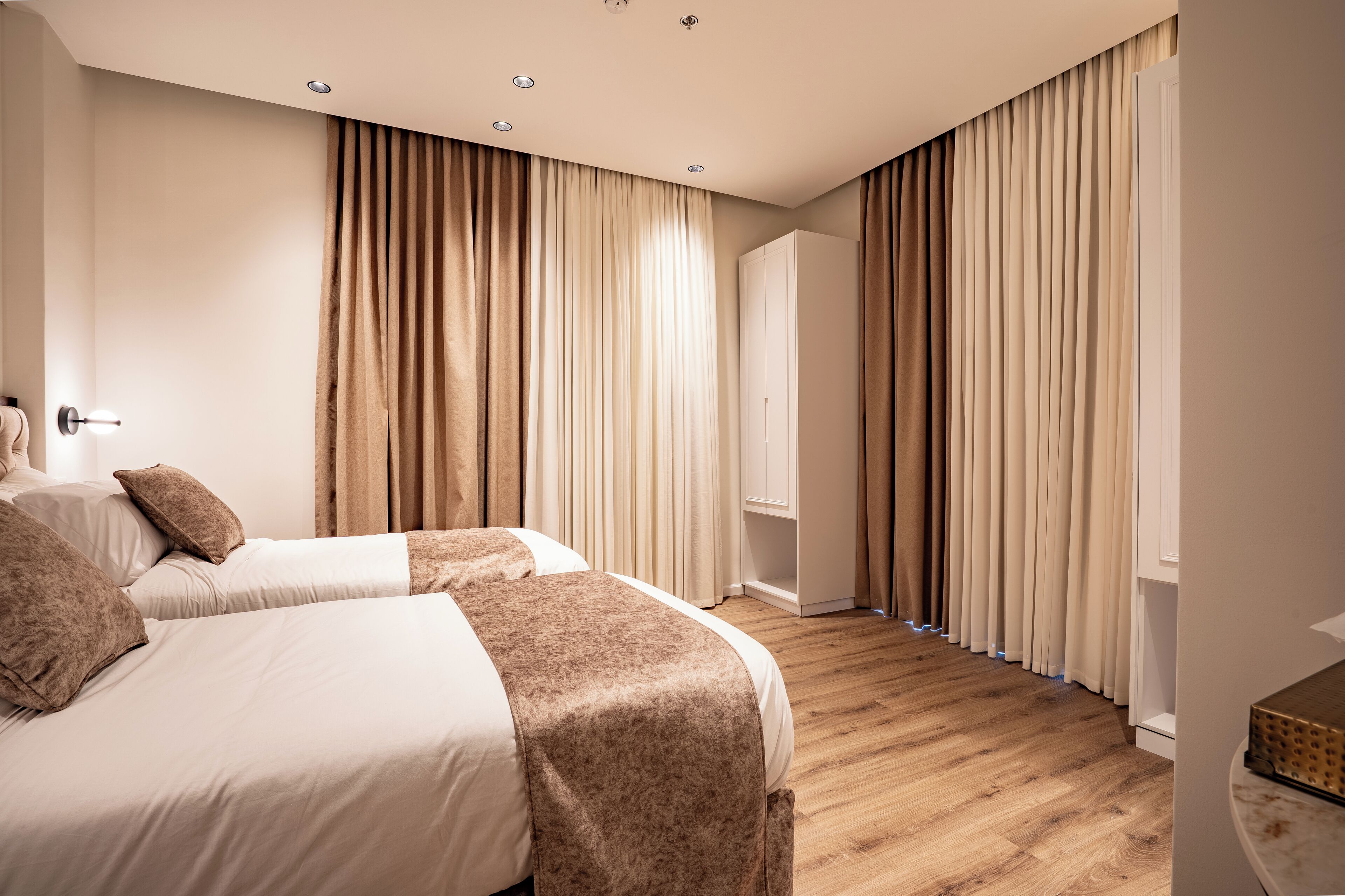 A hotel room with two beds, beige curtains, wooden flooring, and a wardrobe.