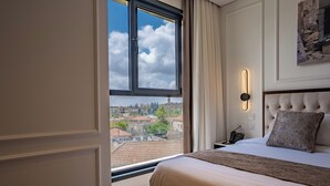 A hotel room with a large window, a bed with a headboard, a bedside table, and a wall-mounted light.
