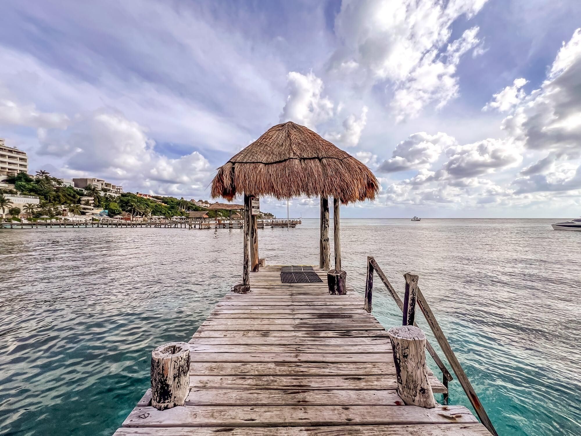 A wooden pier leading to a thatched-roof structure with a view of the sea and distant buildings.