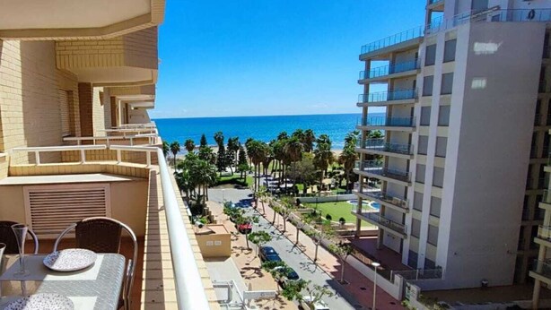 A balcony with a table set for two, overlooking a beach and a parking area with cars.