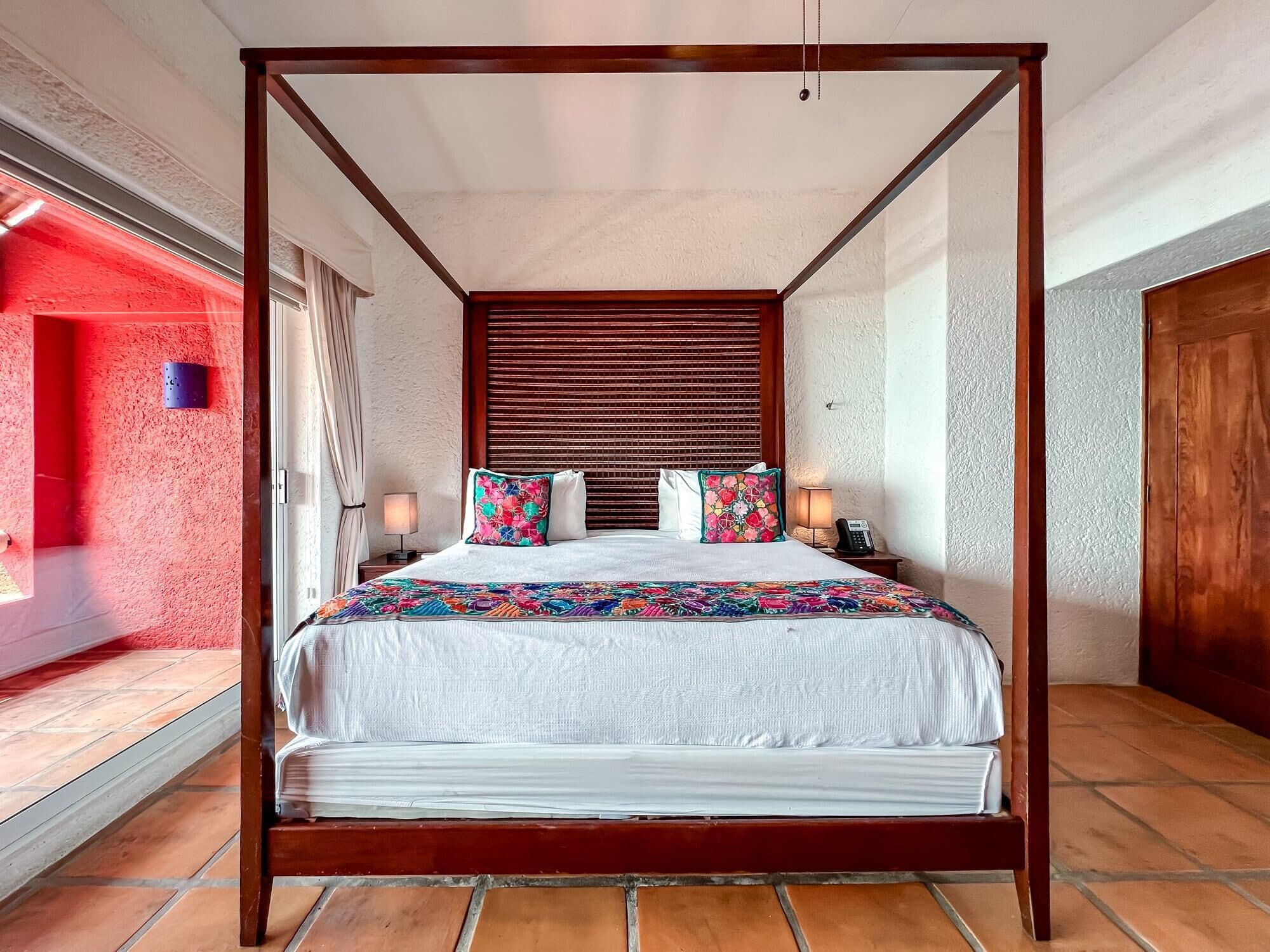 A four-poster bed with a white bedspread and colorful pillows, a bedside table with a lamp, and a telephone.