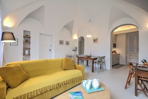 A living room with a yellow sofa, a dining table with chairs, and a kitchen area visible through an archway. - La Dolce Pigna (Sanremo)