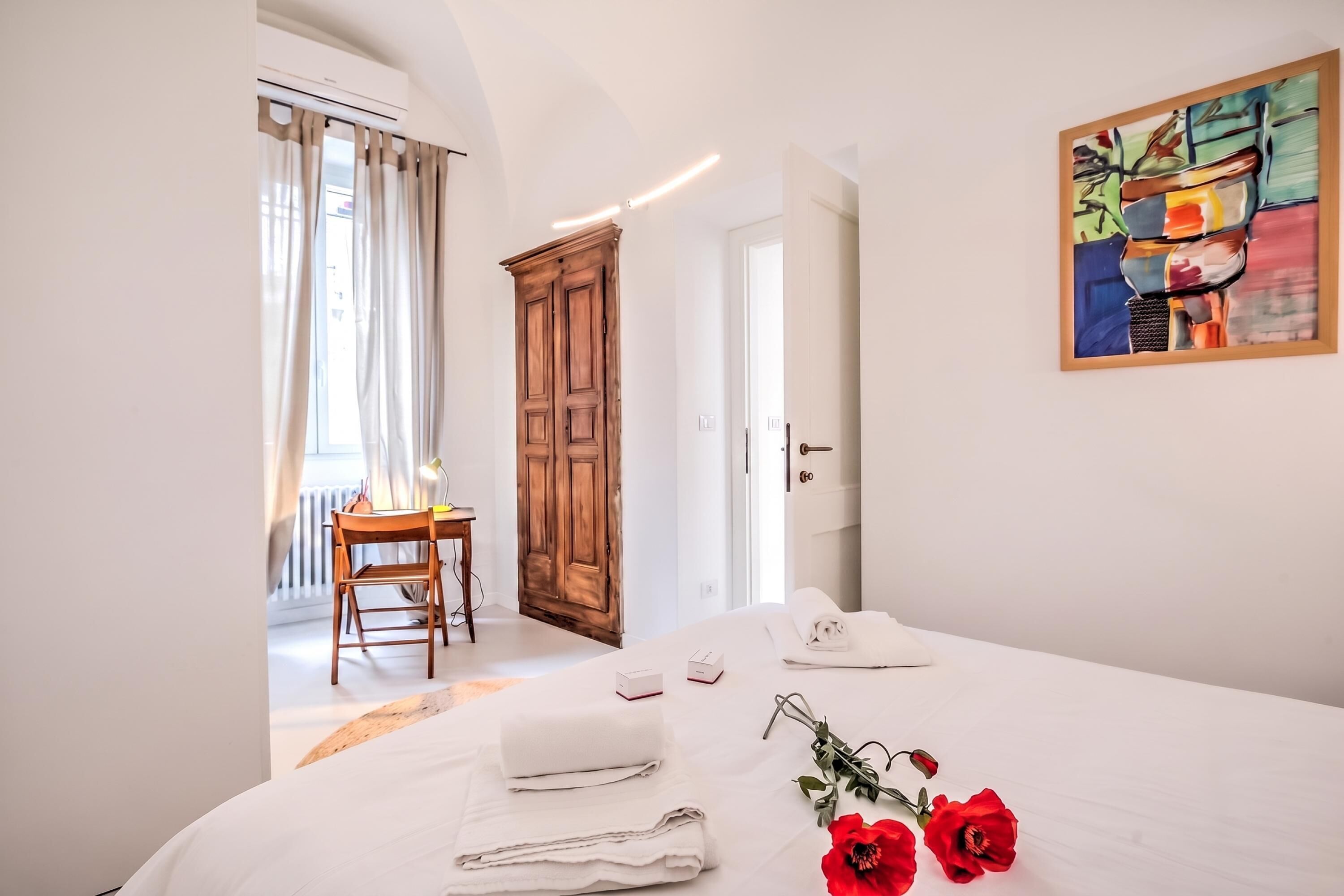 A hotel room with a bed, a desk, a chair, a wardrobe, and a painting on the wall.