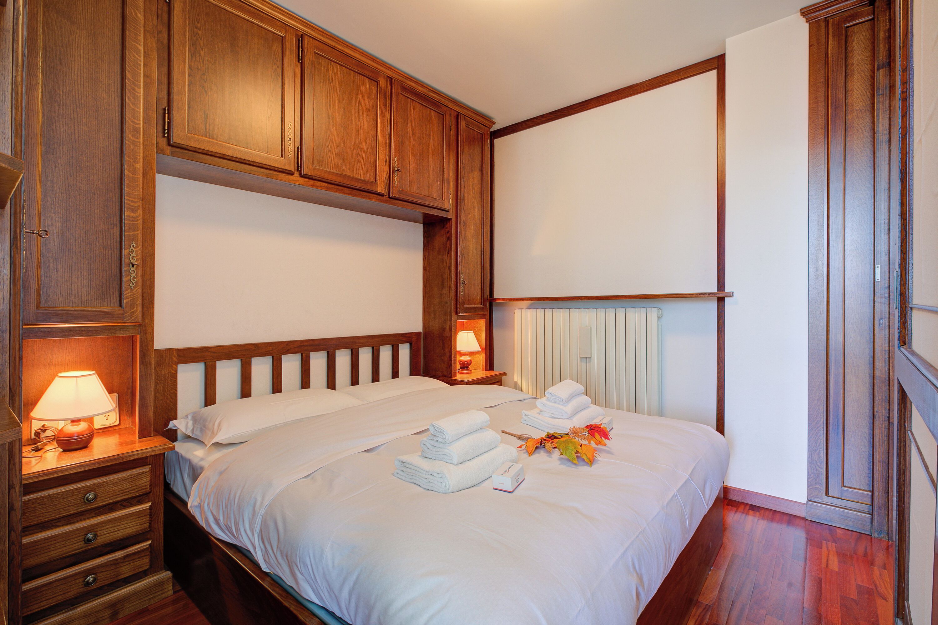 A neatly made bed with white linens and folded towels, wooden headboard, and bedside lamps.