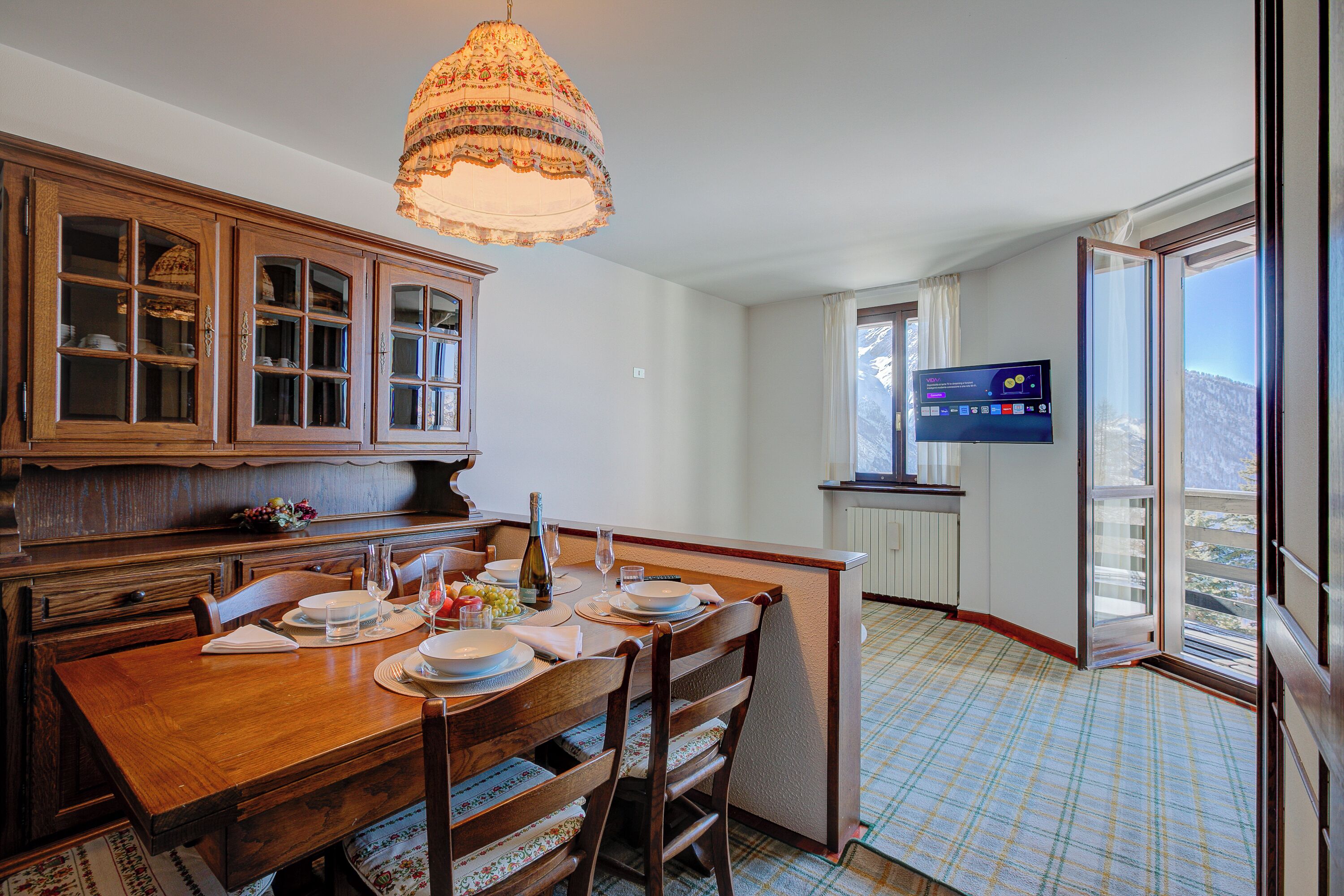 A dining area with a wooden table set for four, a chandelier, and a television mounted on the wall.