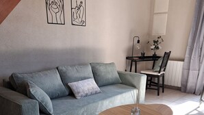 Una sala de estar con un sofá, una mesa redonda de centro y un pequeño reposapiés. Se observa una mesa de comedor con sillas y un radiador al fondo.