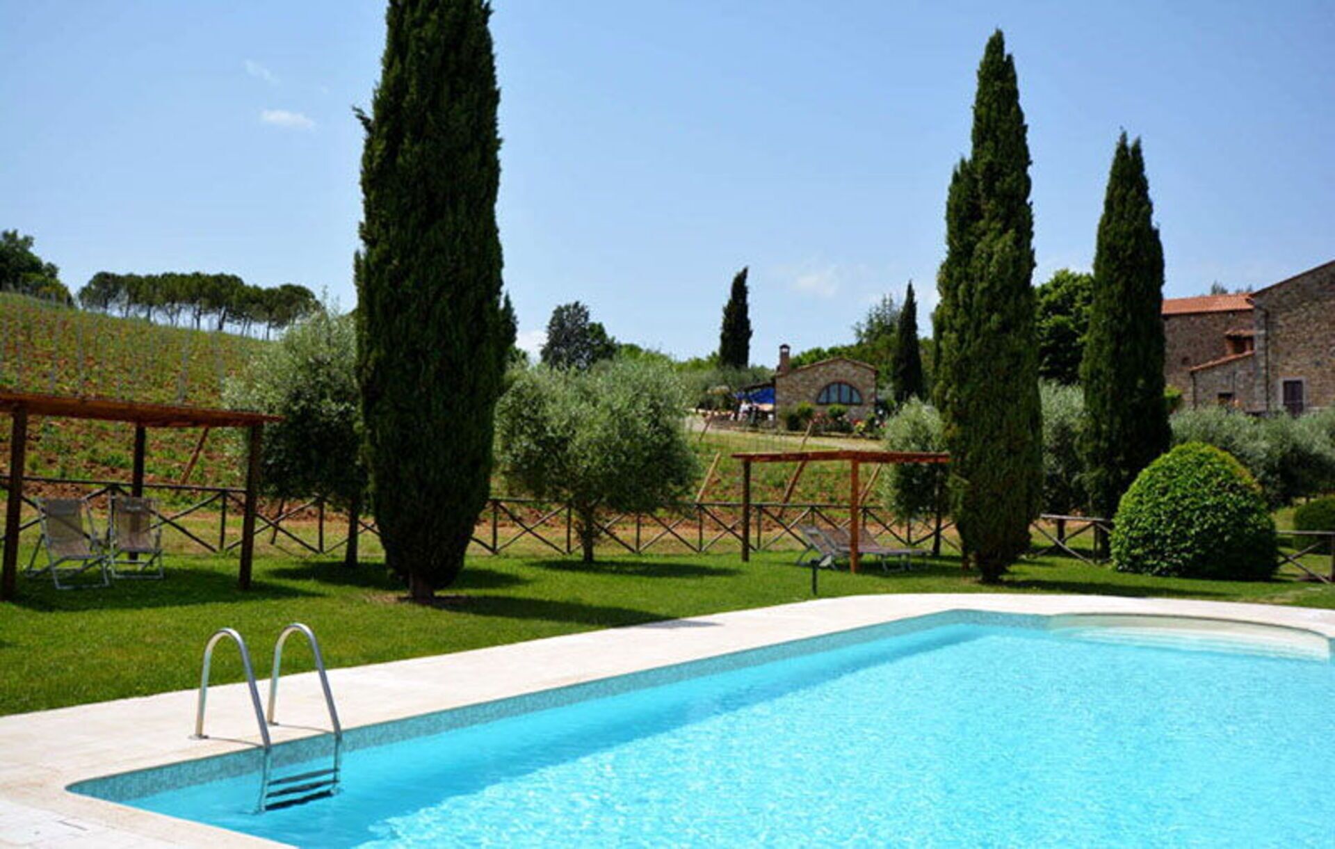 A swimming pool with a wooden deck and metal ladder, surrounded by well-maintained grass and cypress trees.