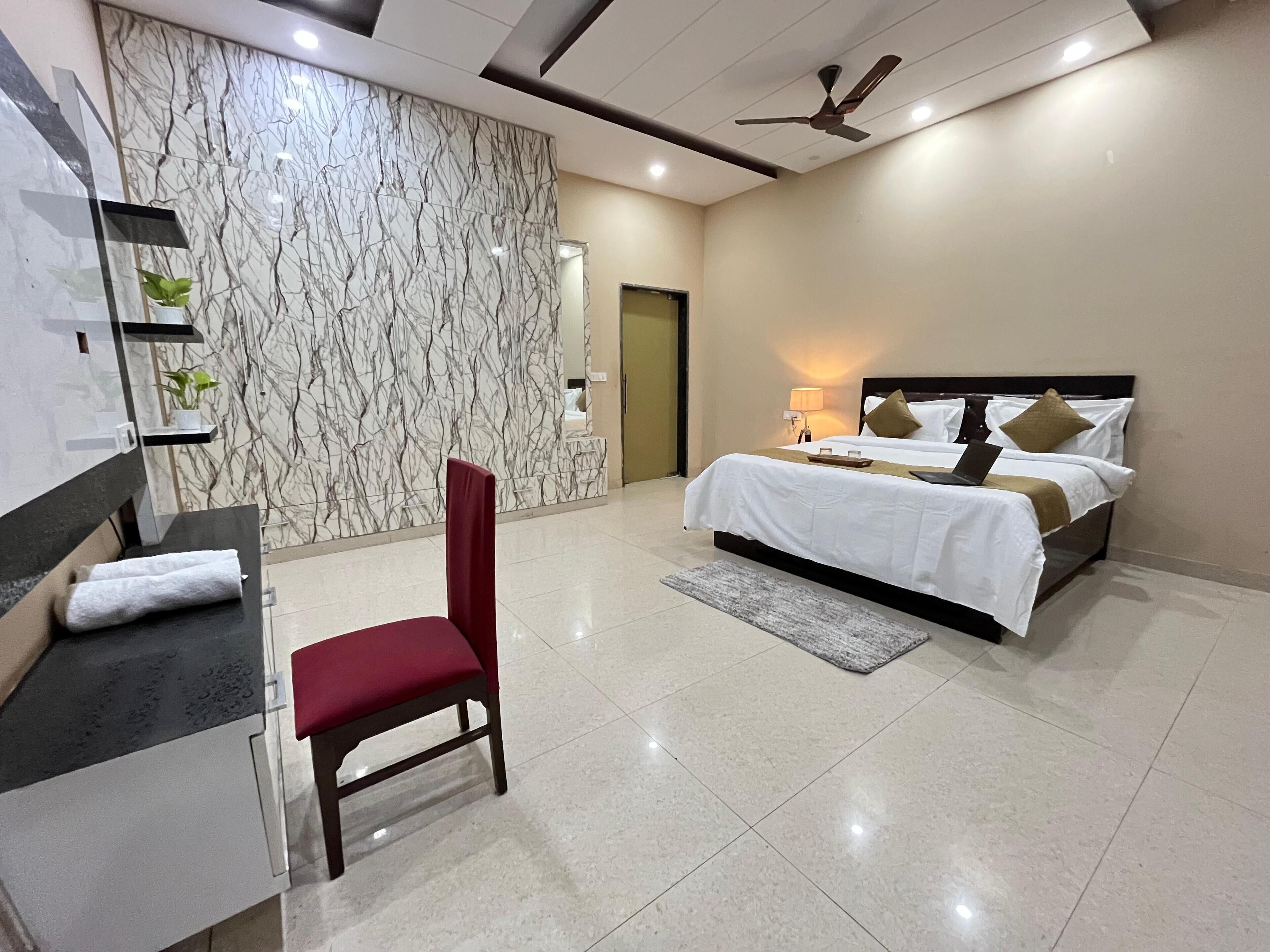 A modern hotel room with a marble wall, a bed with white bedding, a red chair, and a desk with a laptop.