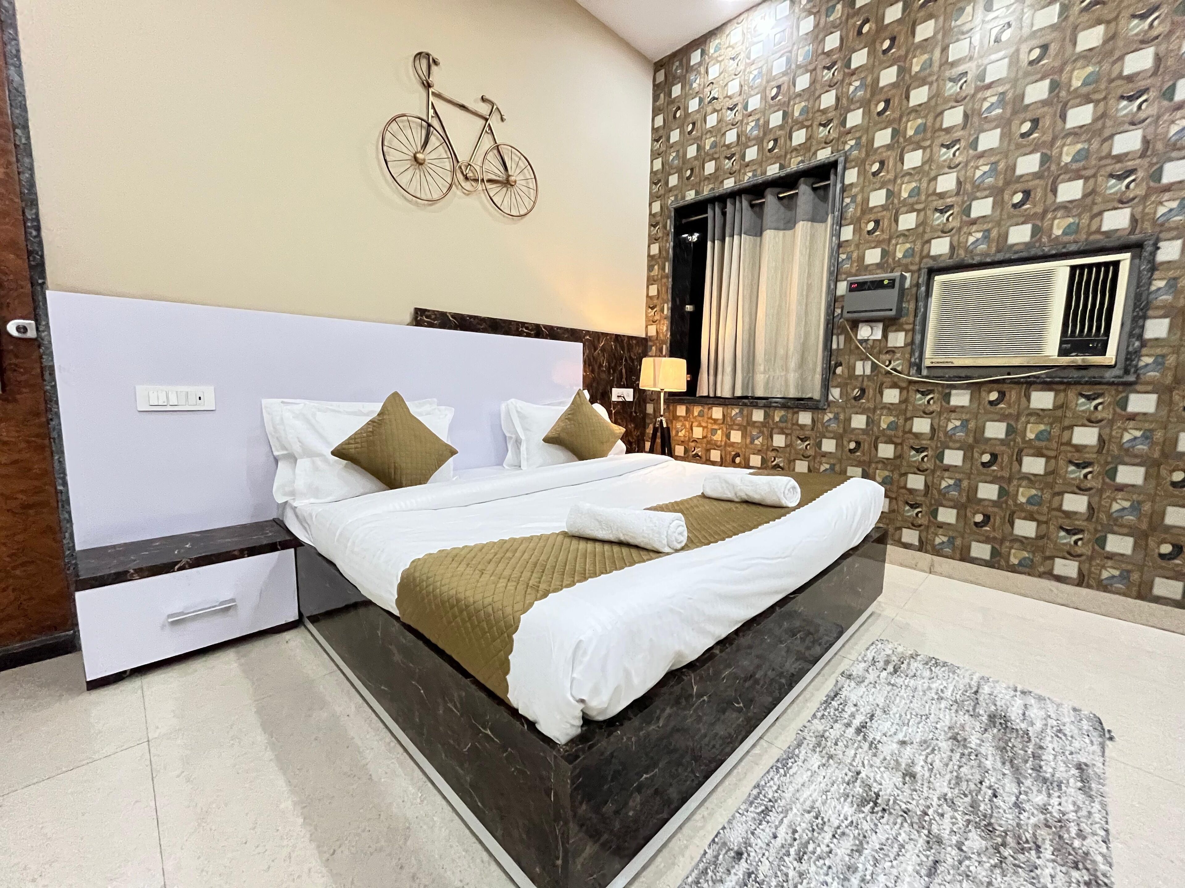 Una camera d'albergo con un letto, un condizionatore d'aria a parete, una parete decorata con una bicicletta e una finestra con tende.