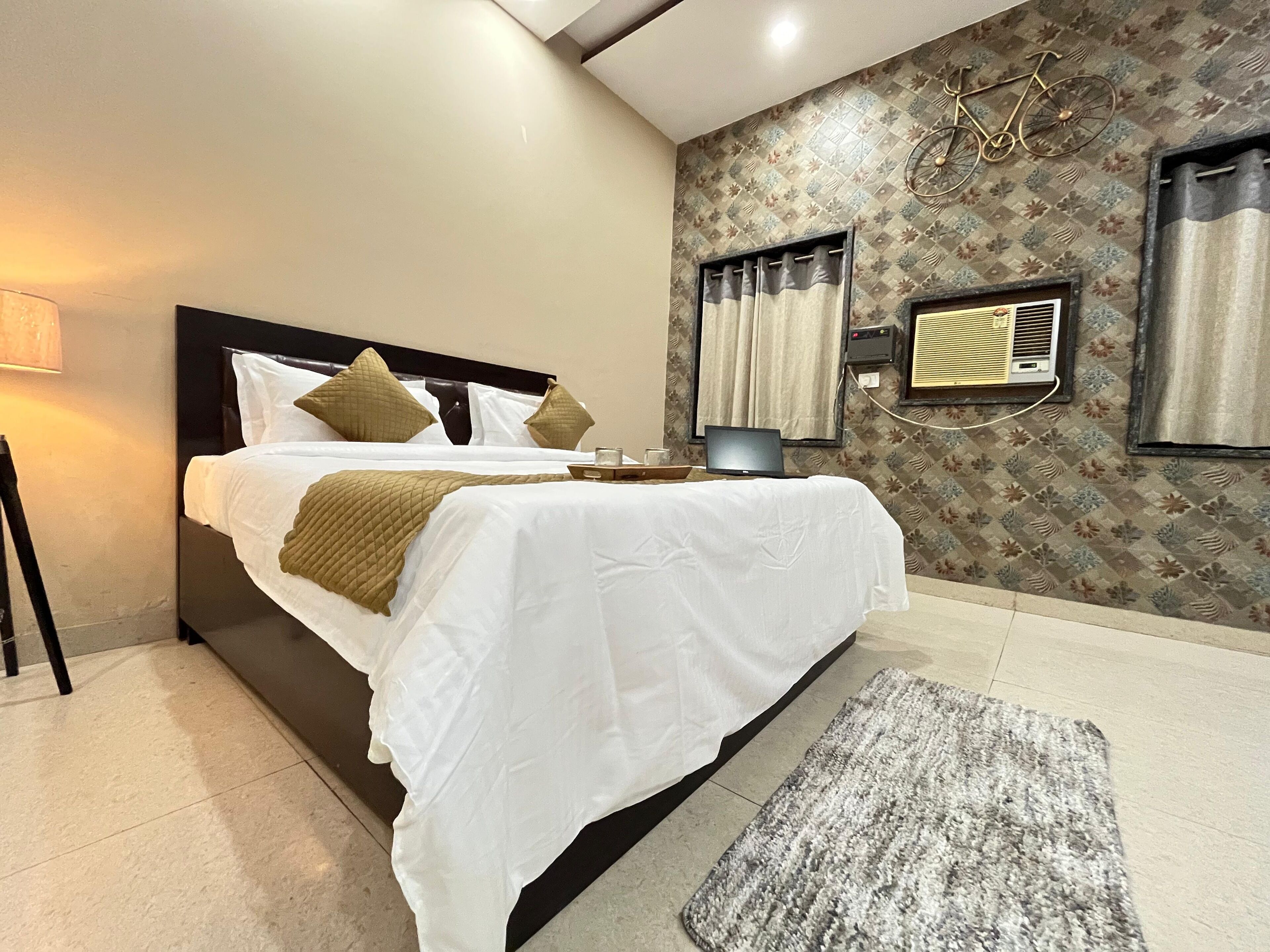 A neatly made bed with white linens and a brown throw, a wall-mounted air conditioner, and a patterned wallpaper.