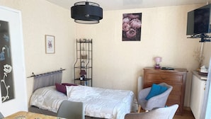 Una habitación pequeña y luminosa con una cama, una silla, una mesa con fruteros, un aparador y un televisor.