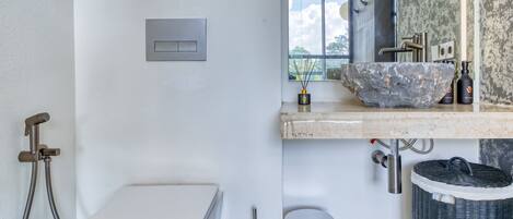 A modern bathroom with a white toilet, a large stone sink, a bidet, and a trash can.