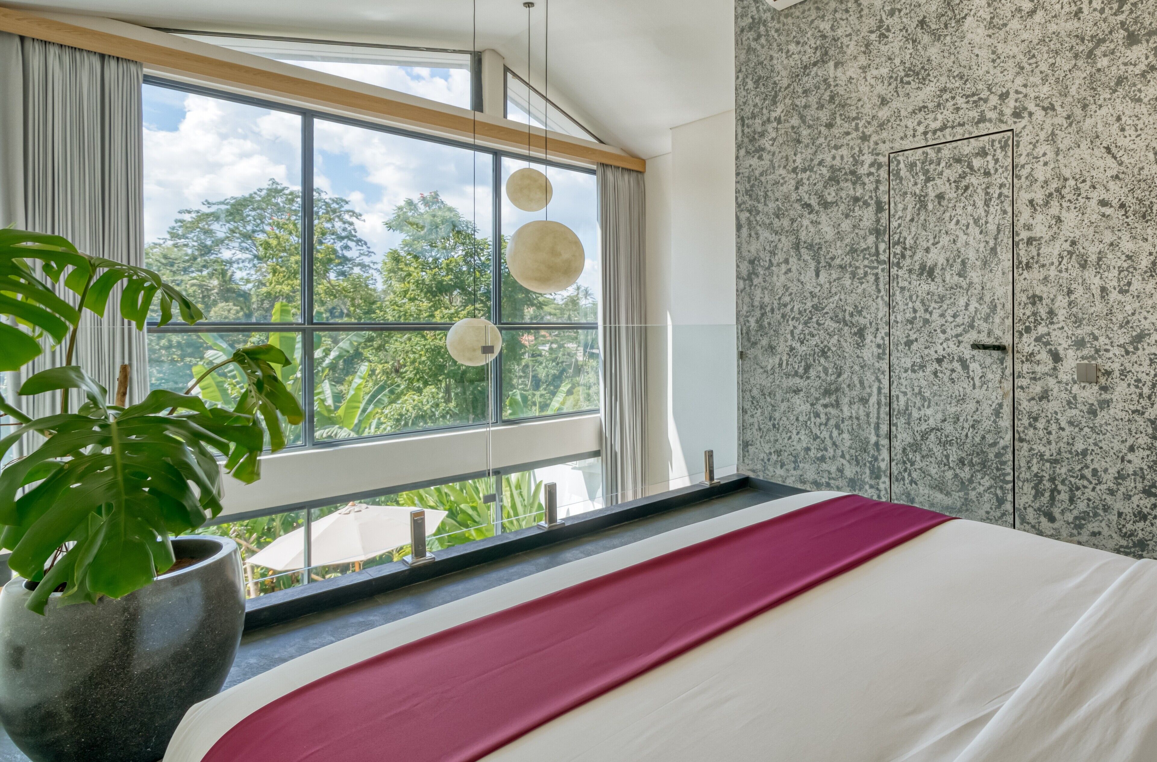 A modern bedroom with a large window, a potted plant, and a bed with a red accent.