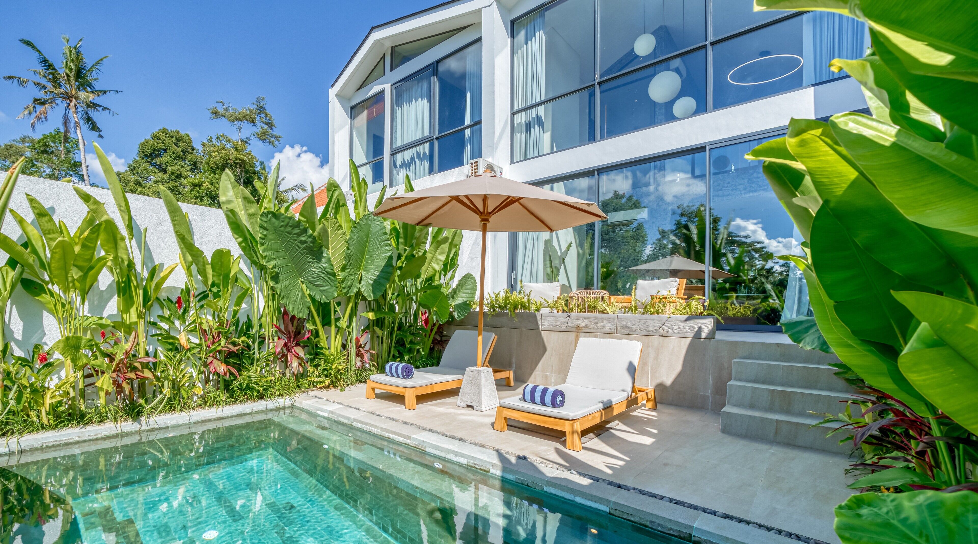 A modern pool area with lounge chairs and an umbrella, surrounded by lush tropical plants.