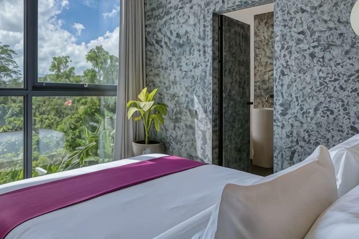 A hotel room with a bed, a window with a view of greenery, and a bathroom visible through an open door.