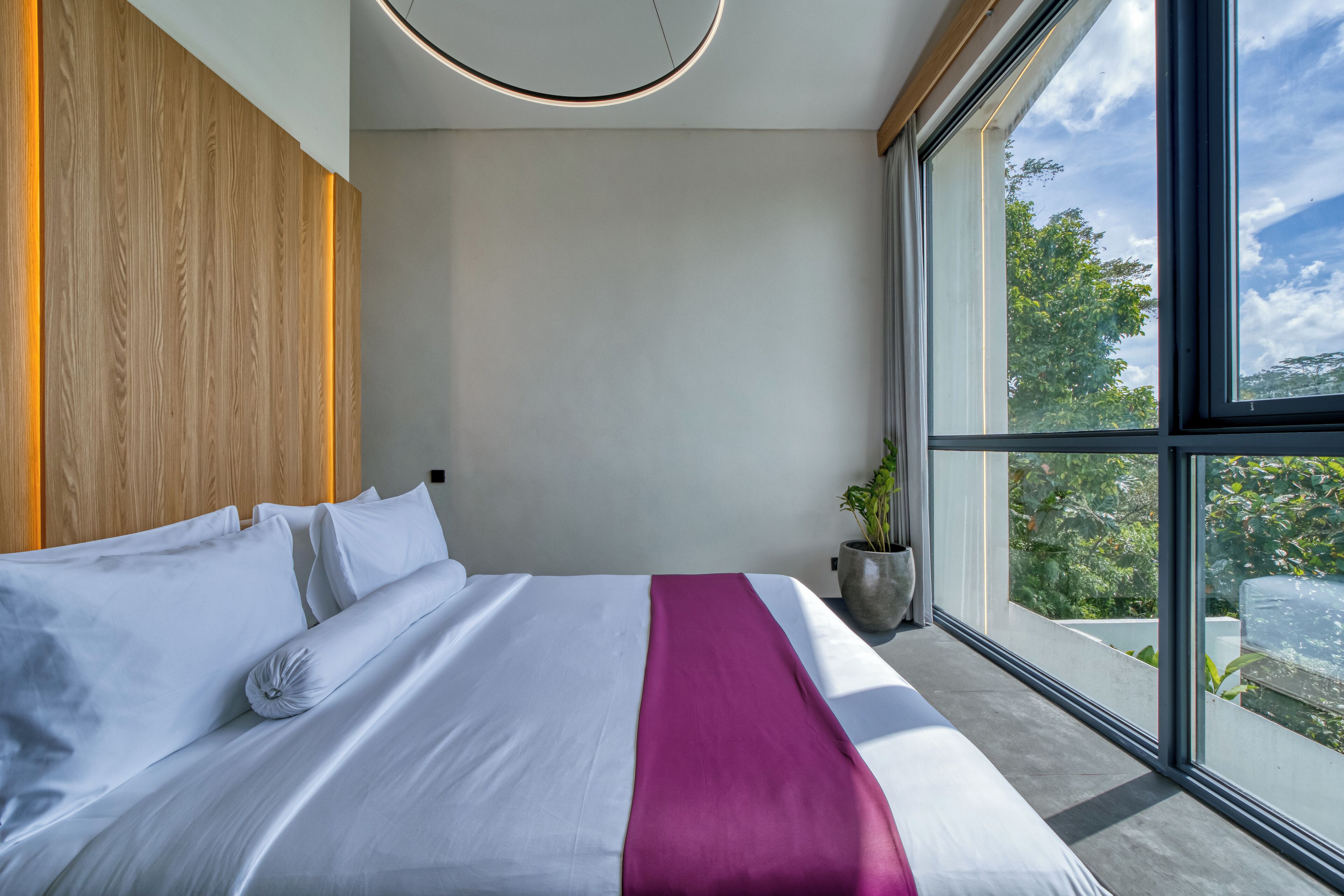 Une chambre d’hôtel moderne avec un grand lit, une tête de lit en bois, une grande fenêtre donnant sur un paysage verdoyant et une plante en pot.