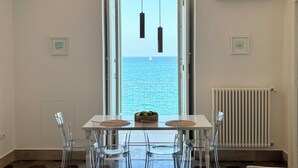 Een eetgedeelte met een tafel en stoelen, uitzicht op de zee door een geopende deur, en een witte radiator aan de muur.
