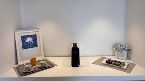 Een minimalistisch hotelkamer met een koffieapparaat, een dienblad met twee kopjes en een mok, een ingelijst kunstwerk en een kleine vaas met bloemen.