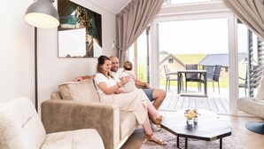 En hyggelig stue med en sofa, et sofabord med blomster og et stort vindue med gardiner.