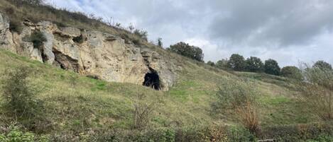 A rocky hillside with a cave entrance, a dirt path, and sparse vegetation.