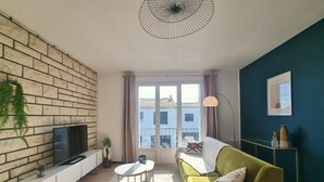 A living room with a TV on a white console, a green sofa, a round table, and a large window with curtains. - Waterfront 200m from beach (Royan)