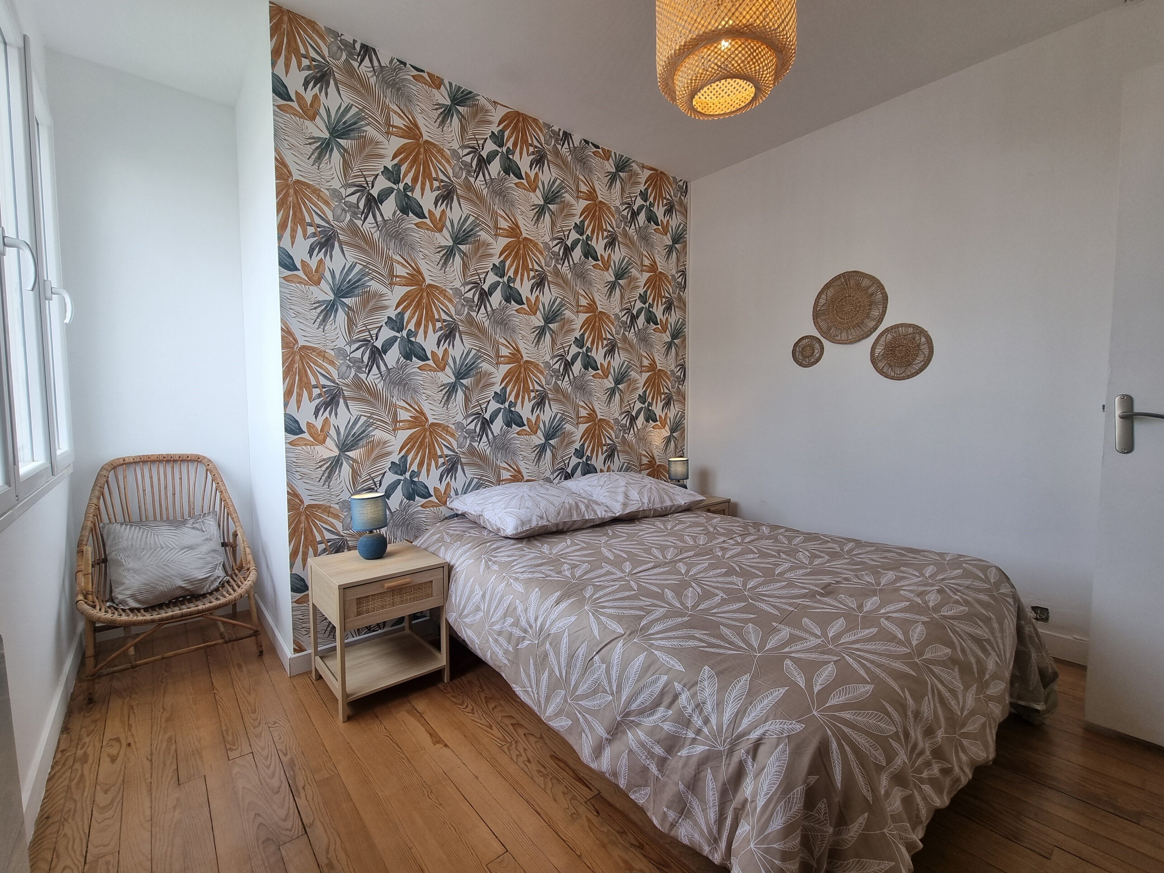 Een slaapkamer met een bed, een nachtkastje, een rieten stoel, een muur met een tropisch bladpatroon en een hanglamp.