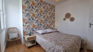 Een slaapkamer met een bed, een nachtkastje, een rieten stoel, een muur met een tropisch bladpatroon en een hanglamp.