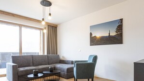 Ein modernes Wohnzimmer mit einem grauen Sofa, zwei runden Couchtischen, einem türkisfarbenen Sessel und einem großen Fenster mit Vorhängen.