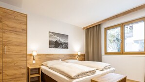 Ein Schlafzimmer mit einem Holzkleiderschrank, einem Bett mit zwei Kissen, einem Nachttisch, einer Wandlampe und einem Fenster mit Vorhängen. An der Wand hängt ein gerahmtes Bild.