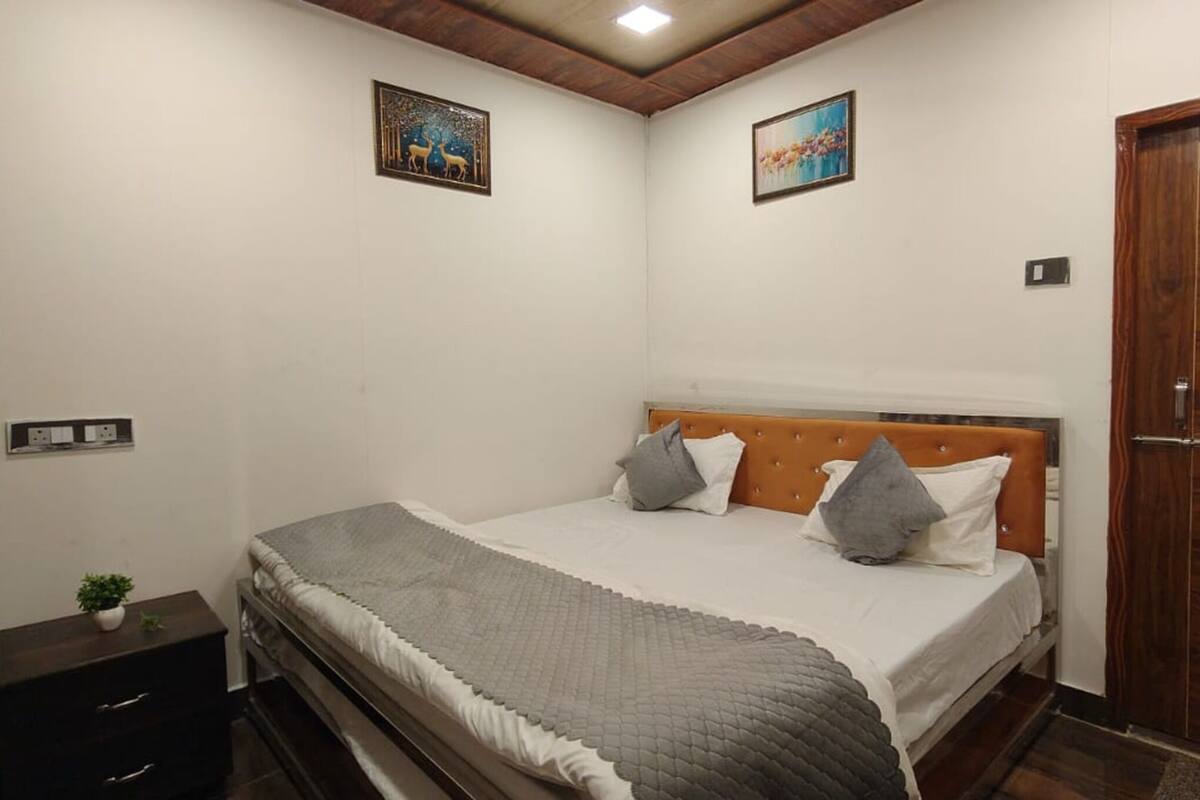 Un dormitorio con cabecera de madera, cama con ropa de cama gris, dos almohadas, una mesita de noche con una planta y una puerta de madera.
