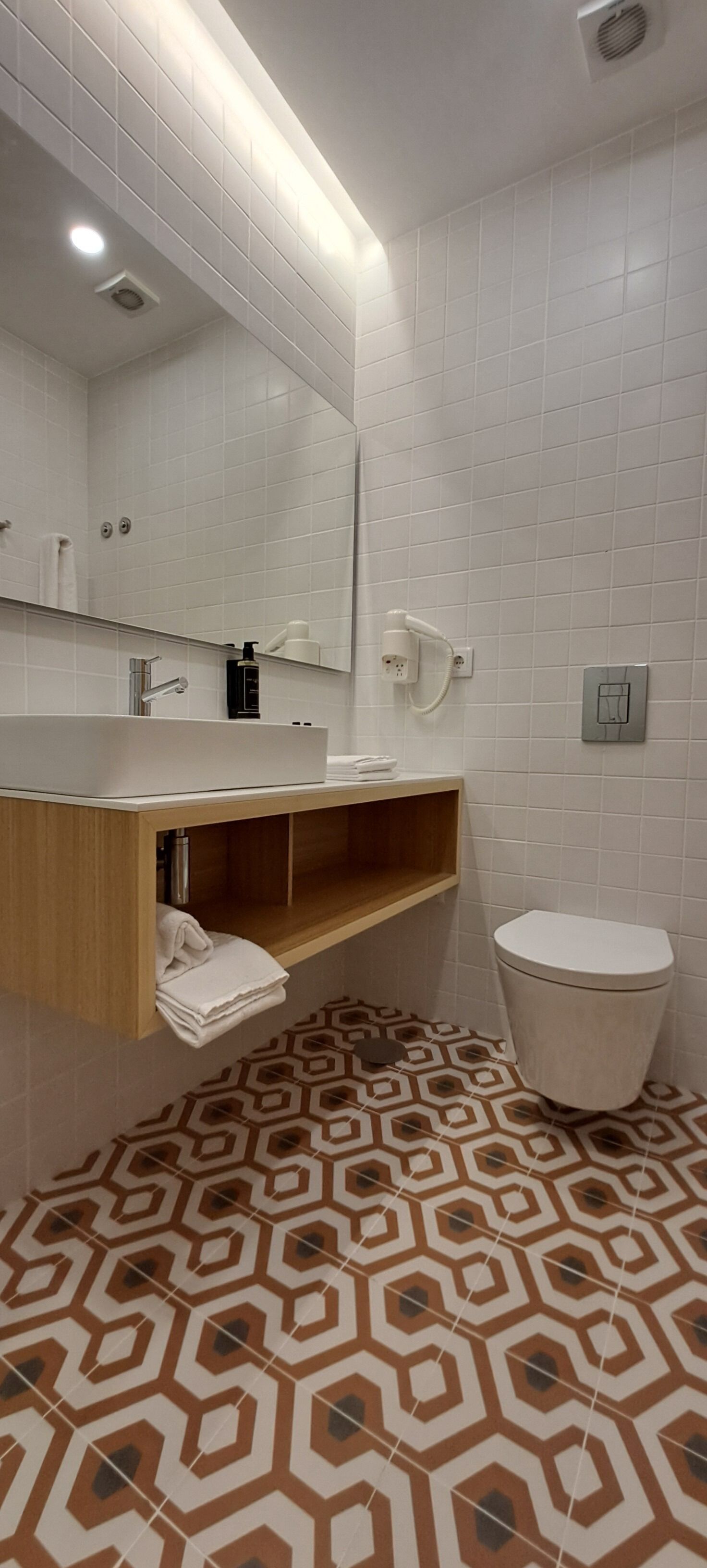 Un baño con inodoro blanco, lavabo con espejo y piso estampado.