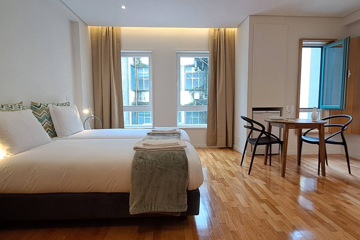 A hotel room with a double bed, a small desk, and a chair. There are two windows with curtains, a wooden floor, and a view of buildings outside.