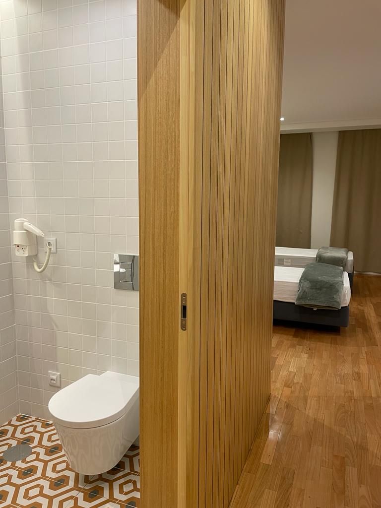 A hotel room with a wooden wardrobe, a white toilet, a patterned floor, and a bed with a blanket.