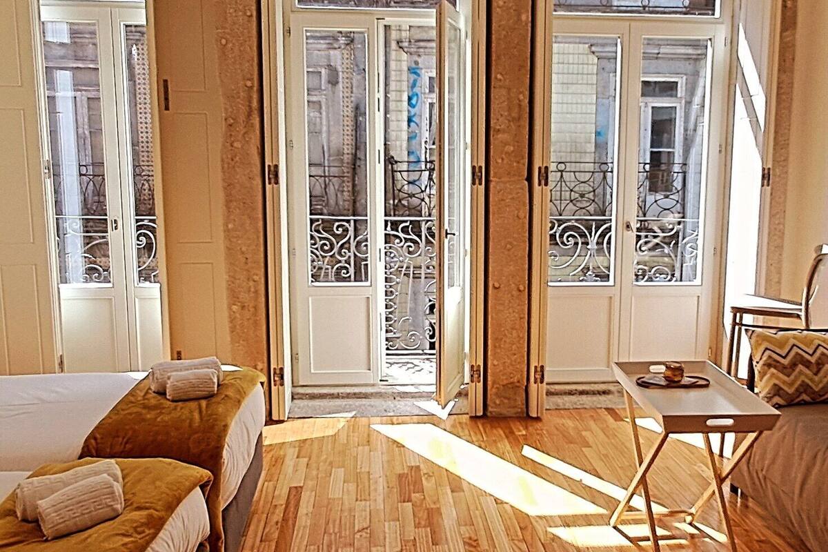 A hotel room with a bed, a small table, and a chair. There are two doors leading to a balcony with a metal railing.