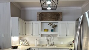 Una cocina con armarios blancos, suelo de madera, un refrigerador de acero inoxidable y una lámpara colgante.