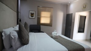 A neatly arranged bedroom with a bed, pillows, a chair, a bedside table, a window with blinds, and a door. - Dikukama Executive Hotel (Tshabong)