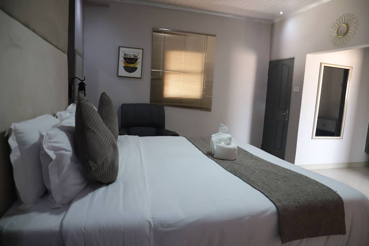 Quarto bem arrumado com cama, travesseiros, uma cadeira, mesa de cabeceira, janela com persianas e uma porta.