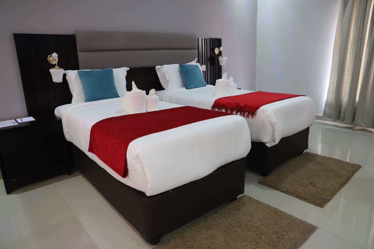 Quarto de hotel moderno com duas camas, cabeceira escura, roupa de cama branca e manta vermelha.