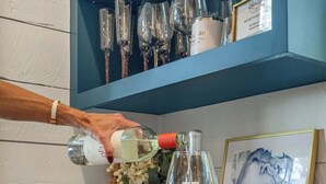 Eine Hand gießt eine klare Flüssigkeit in ein Weinglas auf einer Holzauskidde mit einem blauen Schrank und Regalen darüber, die verschiedene Gläser enthalten.