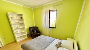 A bedroom with a bed, a chair, a white shelving unit, and a window with curtains. - Apartman Niko (Crikvenica)