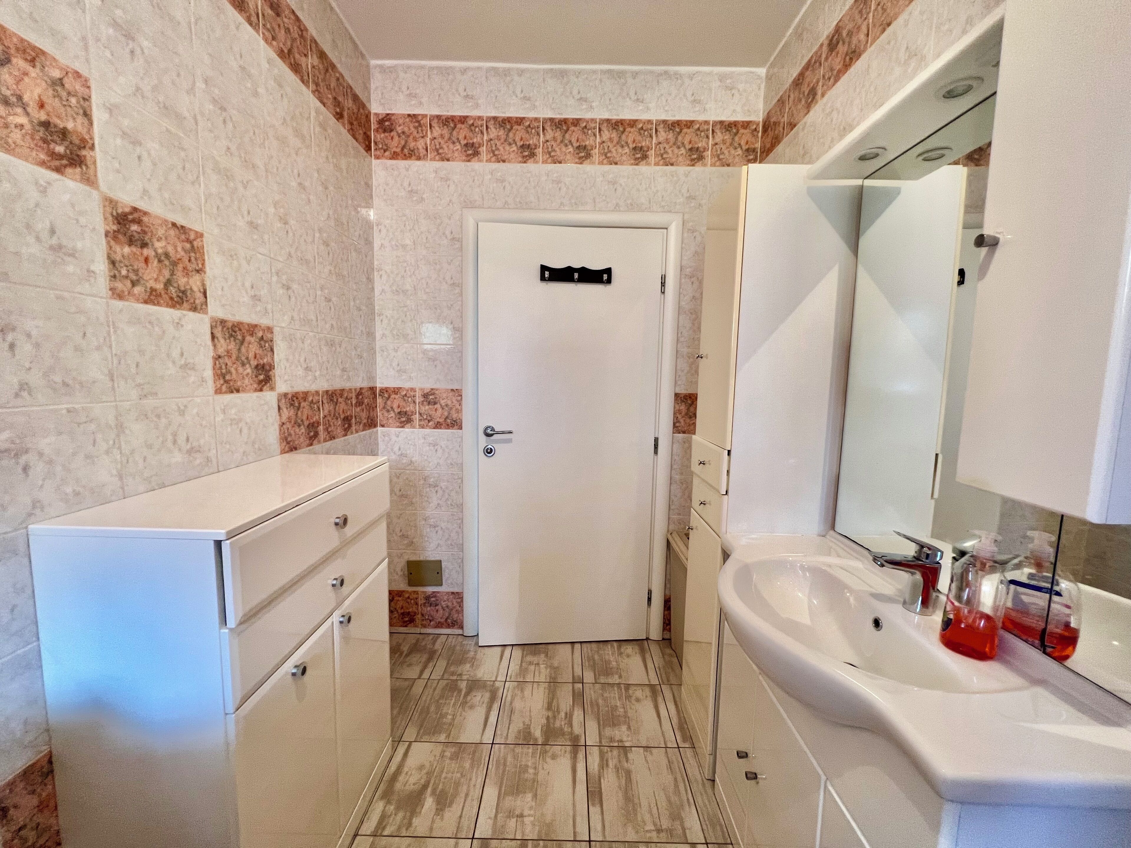 A bathroom with a white door, a sink with a mirror, and a cabinet with drawers.