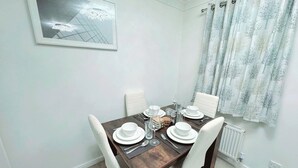 Comedor con una mesa rectangular dispuesta para cuatro personas, sillas blancas, un cuadro en la pared y una ventana con cortinas estampadas.